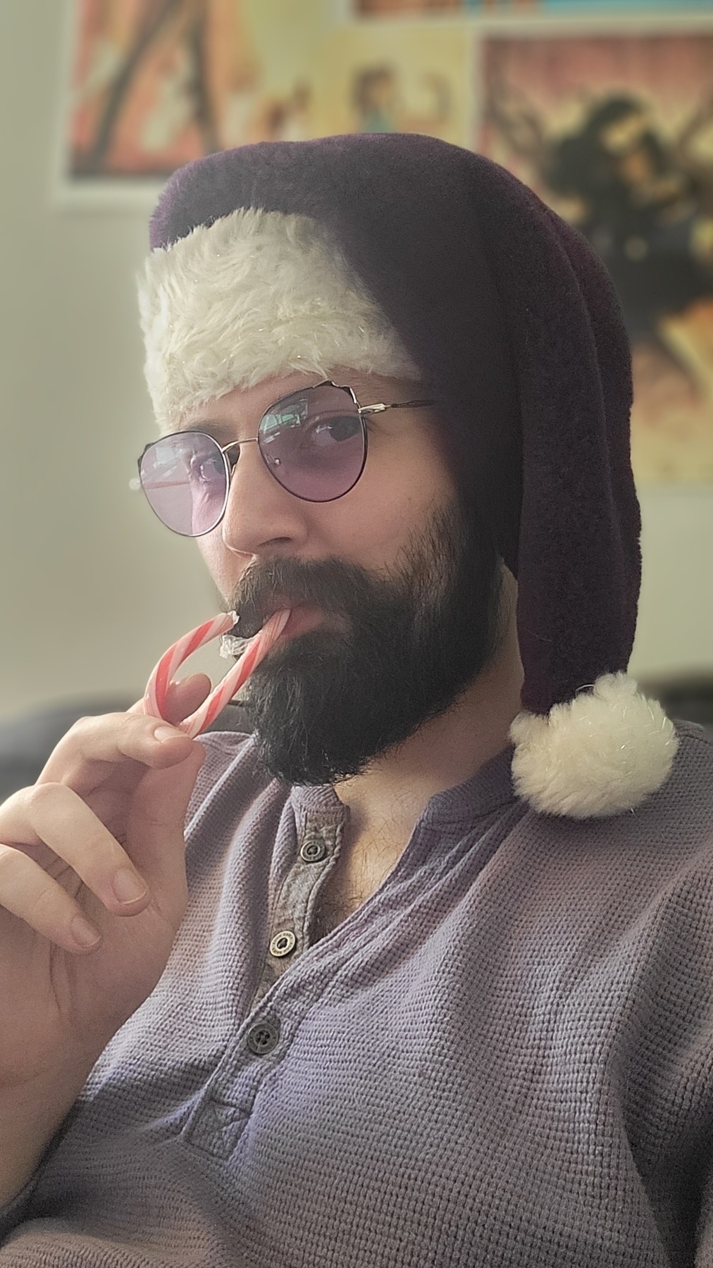 Me (bearded gentleman) smiling at camera while sucking a candy cane, smirking, looking sideways at camera through purple tinted glasses, wearing a purple knit top and a plush purple Santa hat