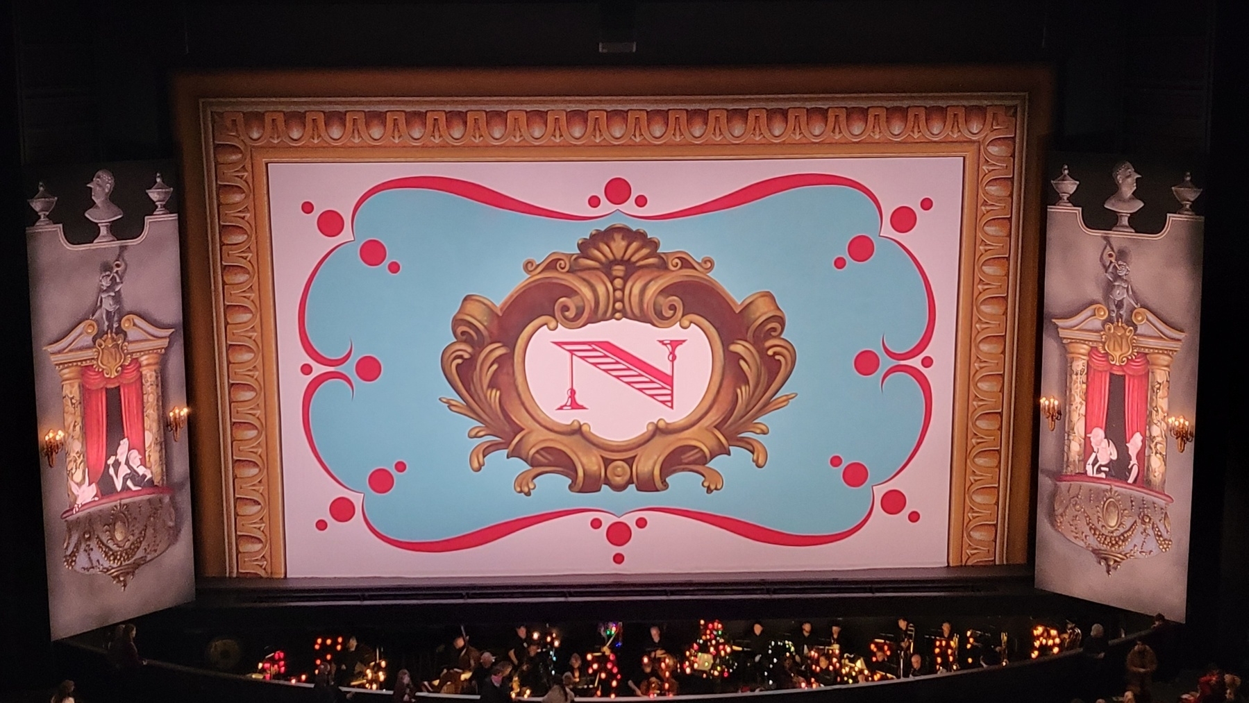 The stage and orchestra pit at the ballet. The dropdown in front of the stage is decorated with an ornate frame and a fancy letter N. There are painted theater boxes on either side. The orchestra pit is full of soft Christmas lights.